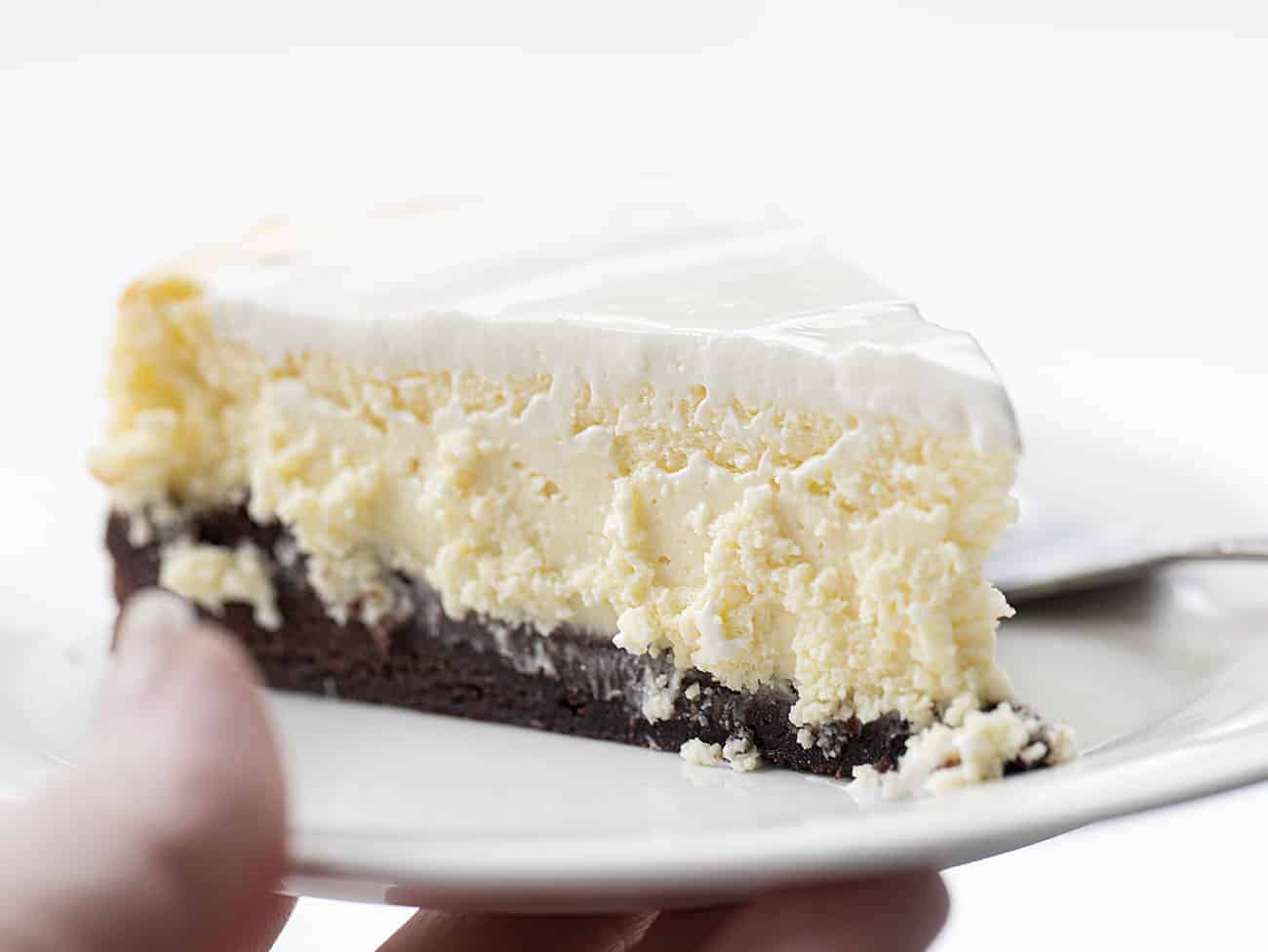 递拿着有果仁巧克力底部的乳酪蛋糕食谱的手白色板材与酸性稀奶油结霜的顶部和叉子休息旁边的叉子
